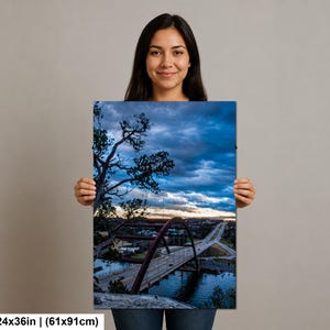 May include: A rectangular wall art print measuring 24 by 36 inches, featuring a landscape photograph of a large arched bridge over a body of water at dusk. The scene includes a dark, cloudy blue sky, silhouetted trees, and a highway with traffic.