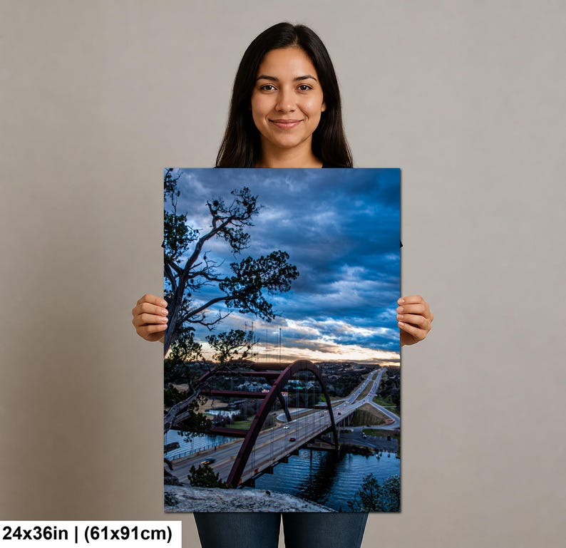 May include: A rectangular wall art print measuring 24 by 36 inches, featuring a landscape photograph of a large arched bridge over a body of water at dusk. The scene includes a dark, cloudy blue sky, silhouetted trees, and a highway with traffic.