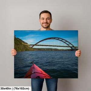 May include: A rectangular wall art print measuring 30 by 40 inches featuring a landscape photograph of a red kayak bow in the foreground on dark blue water, with a large arched bridge spanning across a river under a clear blue sky.
