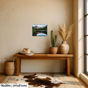 May include: A rectangular wall art print featuring a landscape of mountains, evergreen trees, and a lake with a reflection, displayed above a wooden bench with a beige hat, a potted cactus, and a vase with dried grass on a cowhide rug.