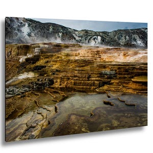 K&ouml;nnte beinhalten: Eine Landschaftsaufnahme der Mammoth Hot Springs mit gestuften Travertinterrassen in Orange-, Braun- und Wei&szlig;t&ouml;nen. Dampf steigt von den mineralhaltigen Becken und kaskadierenden Wasserformationen vor einer Kulisse aus felsigen Bergen auf.