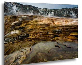 Puede incluir: Una fotografía de paisaje de Mammoth Hot Springs con terrazas de travertino escalonadas en tonos naranja, marrón y blanco. El vapor se eleva de las piscinas ricas en minerales y las formaciones de agua en cascada frente a montañas rocosas.