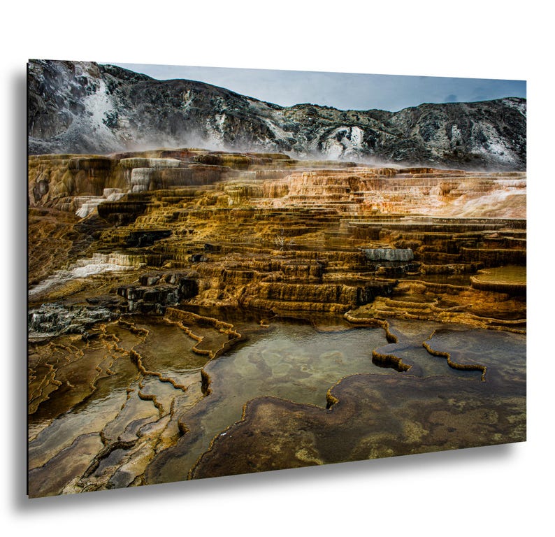 K&ouml;nnte beinhalten: Eine Landschaftsaufnahme der Mammoth Hot Springs mit gestuften Travertinterrassen in Orange-, Braun- und Wei&szlig;t&ouml;nen. Dampf steigt von den mineralhaltigen Becken und kaskadierenden Wasserformationen vor einer Kulisse aus felsigen Bergen auf.
