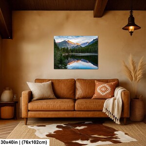 May include: A rectangular wall art print featuring a mountain landscape with a lake, pine trees, and snow-capped peaks at sunrise. The print measures 30 by 40 inches and is displayed above a brown leather sofa with two throw pillows and a blanket.