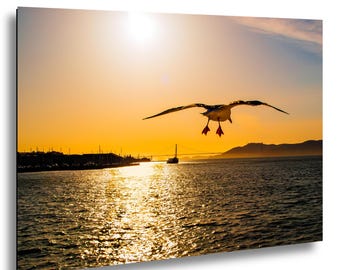May include: A seagull in flight with wings spread wide against a bright golden sunset over a body of water. The sun reflects on the rippling surface, while a distant bridge and landmass are visible on the horizon under a clear orange sky.