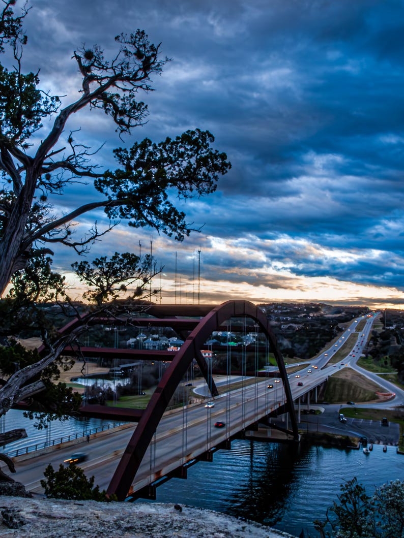 May include: A large steel arch bridge spans a river during twilight, featuring a prominent reddish-brown metal structure with vertical suspension cables. A highway with moving vehicles runs across the bridge under a dramatic, dark blue cloudy sky.