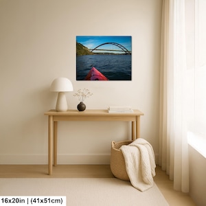 May include: A rectangular wall art print measuring 16 by 20 inches, featuring a landscape photograph of a red kayak bow on dark blue water, looking toward a large arched bridge under a bright blue sky with light clouds, displayed on a wall.