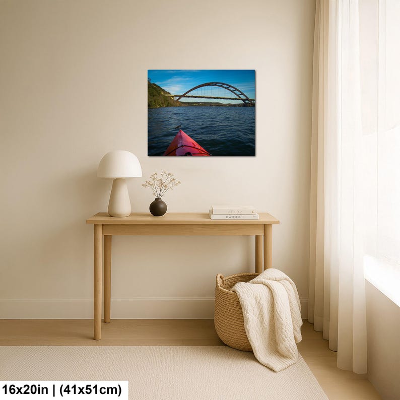 May include: A rectangular wall art print measuring 16 by 20 inches, featuring a landscape photograph of a red kayak bow on dark blue water, looking toward a large arched bridge under a bright blue sky with light clouds, displayed on a wall.