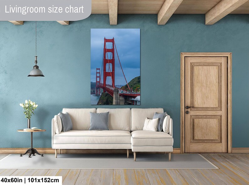 May include: A vertical wall art print of the Golden Gate Bridge in red, displayed above a cream-colored sectional sofa. The print measures 40x60 inches. The scene features the bridge against a cloudy sky, with green hills and water visible below.