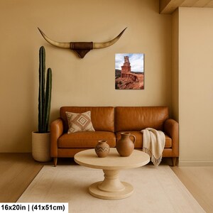 May include: A tan leather sofa with a patterned throw pillow and a cream-colored blanket, a round wooden coffee table with two ceramic vases, a tall cactus in a textured pot, and a wall-mounted longhorn skull above a 16x20 inch desert landscape print.