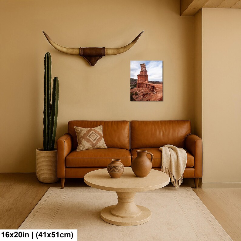 May include: A tan leather sofa with a patterned throw pillow and a cream-colored blanket, a round wooden coffee table with two ceramic vases, a tall cactus in a textured pot, and a wall-mounted longhorn skull above a 16x20 inch desert landscape print.