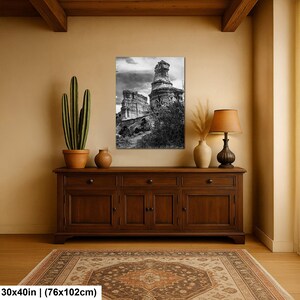 May include: A rectangular wall art print featuring a black and white landscape of tall rock formations under a cloudy sky. The print measures 30 by 40 inches and is displayed above a wooden sideboard with a cactus and a table lamp.