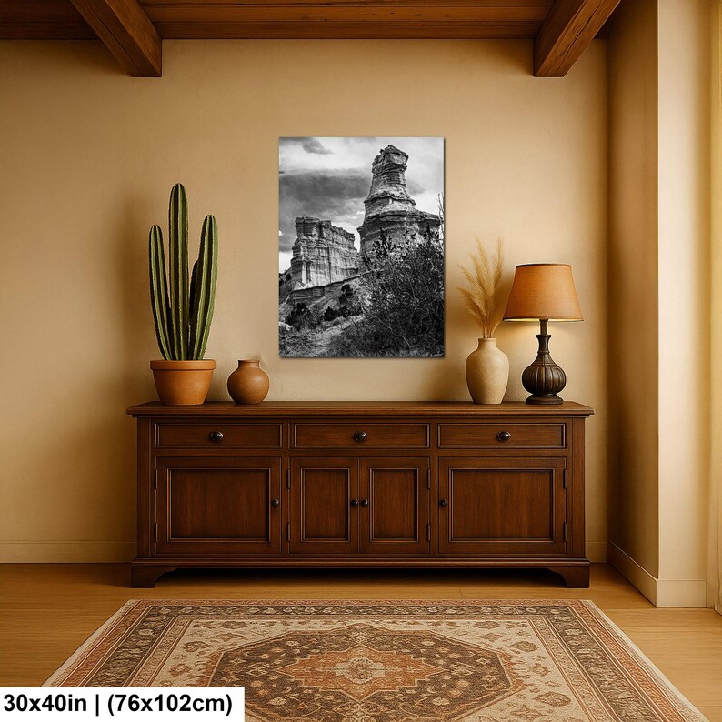 May include: A rectangular wall art print featuring a black and white landscape of tall rock formations under a cloudy sky. The print measures 30 by 40 inches and is displayed above a wooden sideboard with a cactus and a table lamp.