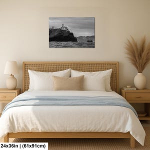 May include: A rectangular black and white wall art print depicting a lighthouse perched on a rocky cliff overlooking the ocean under a cloudy sky. The print measures 24 inches by 36 inches and is displayed above a bed with a wooden headboard.