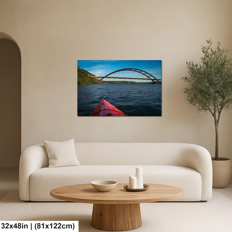 May include: A rectangular wall art print featuring a view from a red kayak on dark blue water, looking toward a large arched bridge under a bright blue sky. The print measures 32 inches by 48 inches and is displayed on a neutral-colored wall.