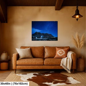 Puede incluir: Una impresi&oacute;n de arte mural rectangular de 76 por 102 cm, que muestra un paisaje nocturno de una cordillera oscura bajo un cielo estrellado azul profundo con una fuente de luz brillante en la base, exhibida sobre un sof&aacute; de cuero marr&oacute;n.