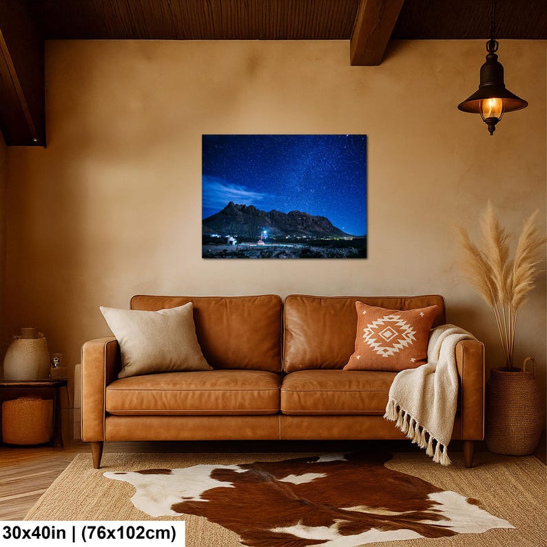 Puede incluir: Una impresi&oacute;n de arte mural rectangular de 76 por 102 cm, que muestra un paisaje nocturno de una cordillera oscura bajo un cielo estrellado azul profundo con una fuente de luz brillante en la base, exhibida sobre un sof&aacute; de cuero marr&oacute;n.