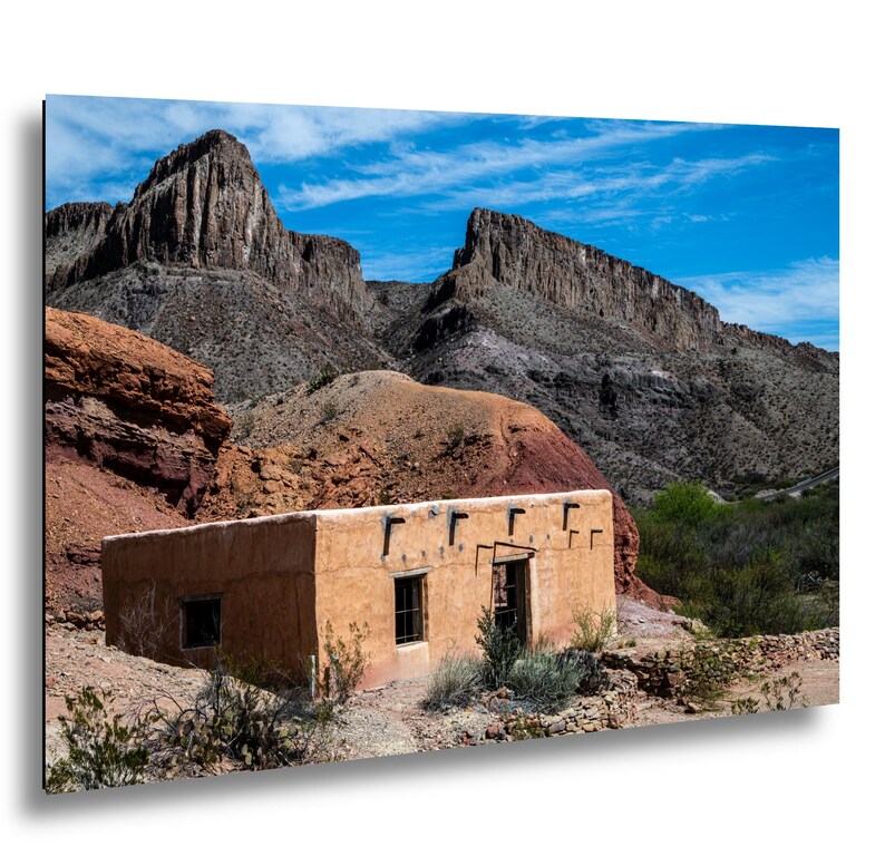 A rectangular adobe building with a flat roof and dark wooden beams protruding from the top, set against a backdrop of rugged, layered brown mountains under a bright blue sky with wispy white clouds and sparse desert vegetation. This is the Contrabando movie set still standing in Big Bend Ranch State Park.