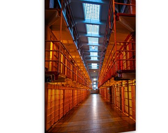May include: A long, narrow prison corridor featuring multiple levels of orange metal cell blocks with vertical bars. The perspective leads toward a bright light at the end of the hallway, with a series of rectangular skylights overhead.