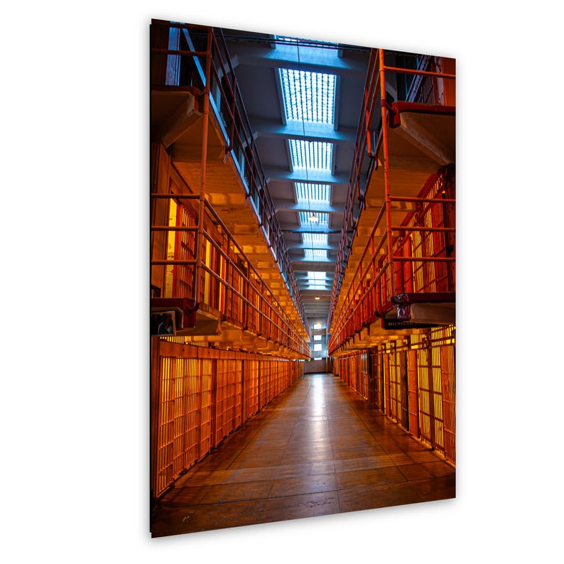 Empty Alcatraz prison corridor in San Francisco, with golden-lit cells, stacked tiers of railings, and overhead skylights converging toward a distant vanishing point. Vertical architectural photograph.
 A long, narrow prison corridor featuring multiple levels of orange metal cell blocks with vertical bars. The perspective leads toward a bright light at the end of the hallway, with a series of rectangular skylights overhead.