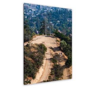 May include: A vertical wall art print featuring a high-angle landscape view of a dirt hiking trail winding up a dry, grassy hill. Two small figures walk along the path toward a hilltop with two tall, thin trees overlooking a sprawling city.