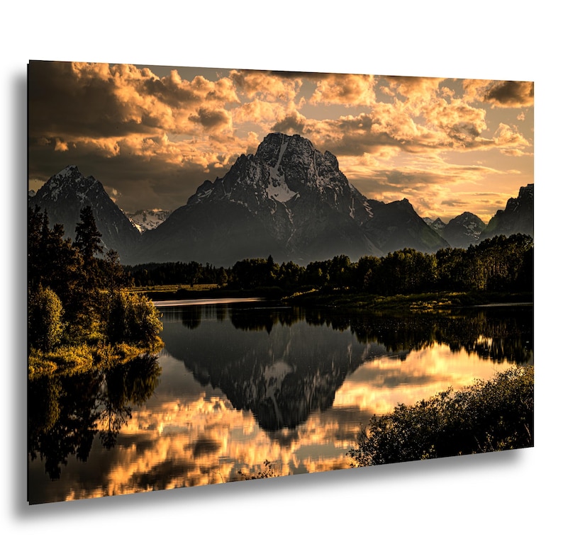 Puede incluir: Un cuadro rectangular que muestra un paisaje de una monta&ntilde;a con picos nevados reflej&aacute;ndose en un lago tranquilo al atardecer. El cielo est&aacute; lleno de nubes doradas y siluetas oscuras de &aacute;rboles bordean la orilla en primer plano.