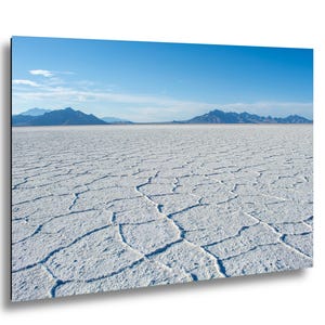 May include: A landscape wall art print featuring the Bonneville Salt Flats with a white, cracked salt crust surface in the foreground. In the distance, dark blue mountain ranges sit under a clear, bright blue sky with light wispy clouds.