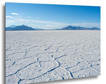 May include: A landscape wall art print featuring the Bonneville Salt Flats with a white, cracked salt crust surface in the foreground. In the distance, dark blue mountain ranges sit under a clear, bright blue sky with light wispy clouds.