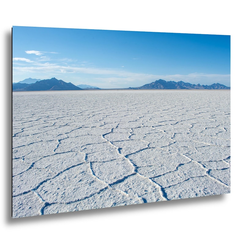 May include: A landscape wall art print featuring the Bonneville Salt Flats with a white, cracked salt crust surface in the foreground. In the distance, dark blue mountain ranges sit under a clear, bright blue sky with light wispy clouds.