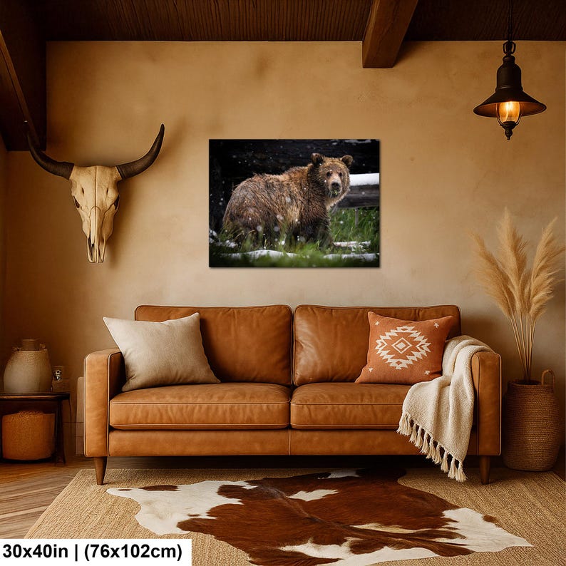 May include: A rectangular wall art print featuring a brown bear standing in a snowy forest, displayed above a brown leather sofa with two throw pillows, a white blanket, a cowhide rug, and a skull mount on the wall. Dimensions: 76x102 cm.