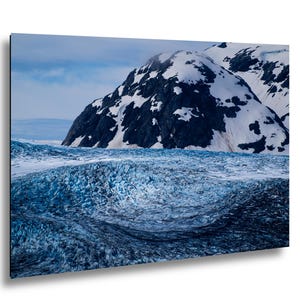 May include: A landscape photograph featuring a vast, textured blue glacier in the foreground with deep crevasses and ice formations. In the background, a large, dark, craggy mountain peak is partially covered in patches of white snow under a cloudy sky.