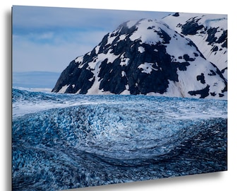 May include: A landscape photograph featuring a vast, textured blue glacier in the foreground with deep crevasses and ice formations. In the background, a large, dark, craggy mountain peak is partially covered in patches of white snow under a cloudy sky.