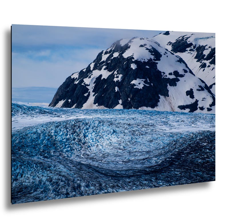 May include: A landscape photograph featuring a vast, textured blue glacier in the foreground with deep crevasses and ice formations. In the background, a large, dark, craggy mountain peak is partially covered in patches of white snow under a cloudy sky.