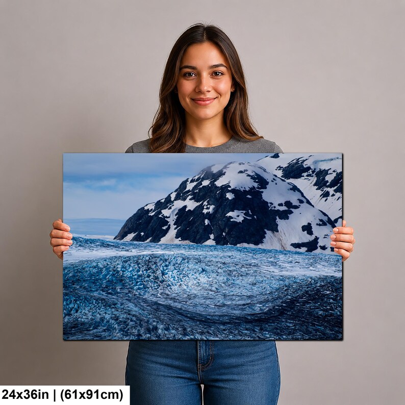 May include: A rectangular wall art print measuring 24x36 inches, featuring a landscape photograph of a deep blue glacier surface in the foreground with snow-capped dark mountain peaks rising in the background under a cloudy sky.