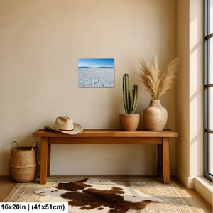 May include: A rectangular wall art print featuring a landscape of a white cracked salt flat under a bright blue sky with distant mountains. The print measures 16 inches by 20 inches and is displayed on a plain wall above a wooden bench.