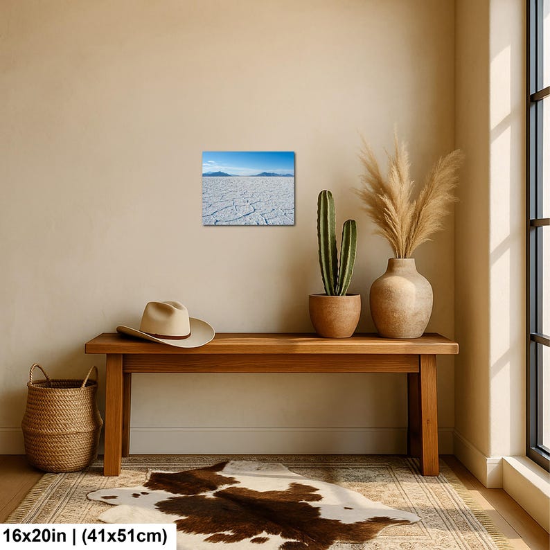 May include: A rectangular wall art print featuring a landscape of a white cracked salt flat under a bright blue sky with distant mountains. The print measures 16 inches by 20 inches and is displayed on a plain wall above a wooden bench.