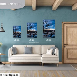 May include: A living room size chart displaying three vertical landscape prints of a bridge at sunset, labeled with dimensions 16" x 24", 20" x 30", and 24" x 36". The prints are shown hanging above a cream-colored sectional sofa in a room.