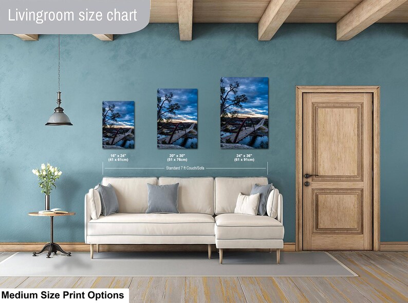 May include: A living room size chart displaying three vertical landscape prints of a bridge at sunset, labeled with dimensions 16" x 24", 20" x 30", and 24" x 36". The prints are shown hanging above a cream-colored sectional sofa in a room.