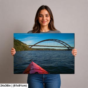 May include: A rectangular wall art print measuring 24x36 inches, featuring a landscape photograph of a red kayak bow in the foreground on dark blue water, with a large arched bridge spanning across the background under a clear blue sky.