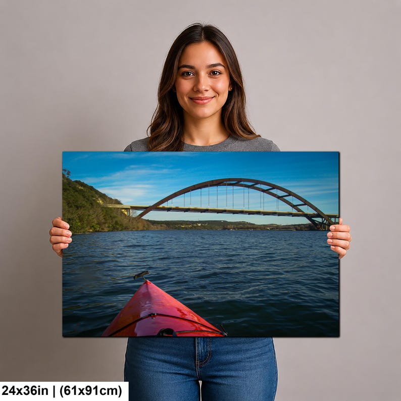 May include: A rectangular wall art print measuring 24x36 inches, featuring a landscape photograph of a red kayak bow in the foreground on dark blue water, with a large arched bridge spanning across the background under a clear blue sky.