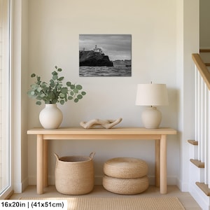 May include: A black and white wall art print depicting a lighthouse perched on a rocky cliff overlooking the ocean under a cloudy sky. The rectangular print measures 16 inches by 20 inches and is displayed in a home interior setting.