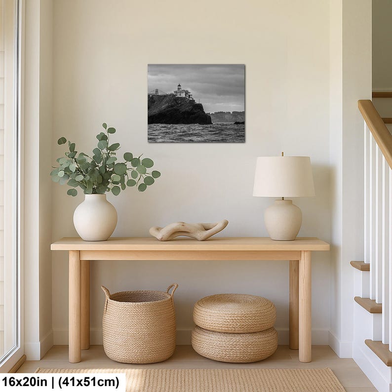 May include: A black and white wall art print depicting a lighthouse perched on a rocky cliff overlooking the ocean under a cloudy sky. The rectangular print measures 16 inches by 20 inches and is displayed in a home interior setting.