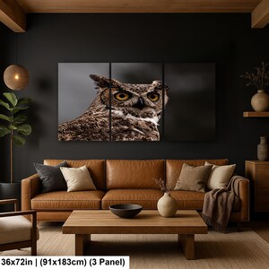 May include: A three-panel wall art set featuring a close-up photograph of a great horned owl with yellow eyes and brown patterned feathers. The artwork measures 36 by 72 inches and is displayed as a triptych on a dark wall above a leather sofa.