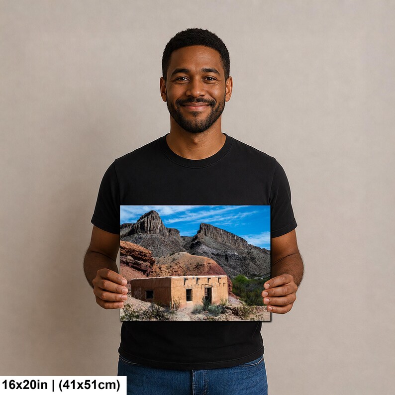 May include: A rectangular wall art print measuring 16x20 inches, featuring a landscape photograph of a tan adobe building situated at the base of rugged, dark rocky mountains under a bright blue sky with scattered clouds.