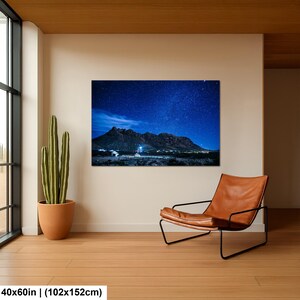 Puede incluir: Una impresi&oacute;n de arte mural rectangular de 102 por 152 cm, que muestra un paisaje nocturno de una cordillera oscura bajo un cielo estrellado azul profundo con una peque&ntilde;a estructura iluminada en la base, sobre una pared color crema.