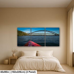 May include: A three-panel wall art set depicting a red kayak bow on dark blue water facing a large arched bridge under a clear blue sky. The artwork measures 40 by 90 inches and features a landscape view of a river scene with a bridge span.