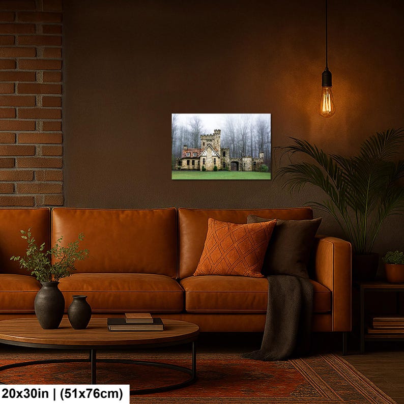 Op de afbeelding: Een rechthoekige wandkunstprint met een stenen kasteel met een toren en een zadeldak, tegen een achtergrond van mistige bomen. De print meet 51 bij 76 cm en hangt aan een donkere muur boven een leren bank.