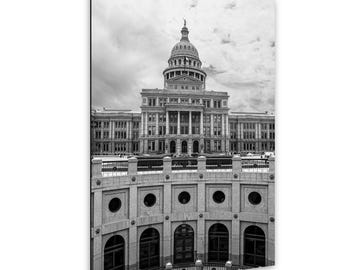 May include: A black and white photograph of the Texas State Capitol building in Austin, featuring its prominent central dome and neoclassical architecture, viewed from an elevated position above a curved stone wall with arched windows.