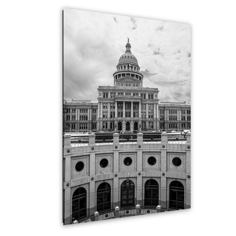 May include: A black and white photograph of the Texas State Capitol building in Austin, featuring its prominent central dome and neoclassical architecture, viewed from an elevated position above a curved stone wall with arched windows.