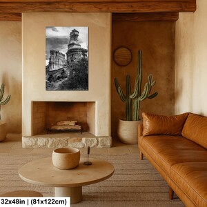 May include: A black and white wall art print depicting a tall, weathered rock formation under a cloudy sky. The rectangular print measures 32 inches by 122 inches and is displayed above a fireplace in a room with a brown leather sofa and cactus.